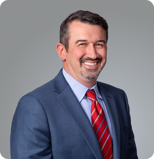 A man with short dark hair, a mustache, and beard is wearing a blue suit, light blue shirt, and red striped tie. He is smiling and posing in front of a plain gray background.