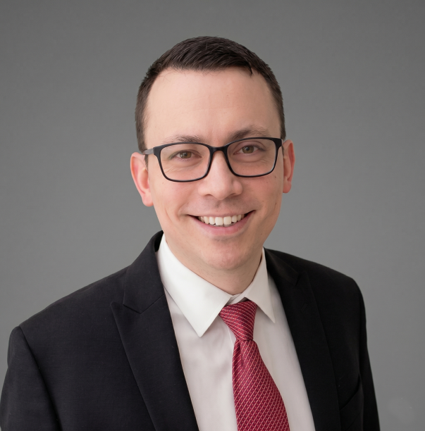 A man wearing glasses, a dark suit, white shirt, and red tie, smiles at the camera in front of a plain gray background.