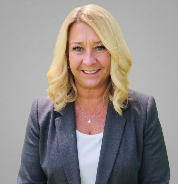 A woman with shoulder-length blonde hair, wearing a gray blazer over a white top, smiles at the camera against a plain light gray background.