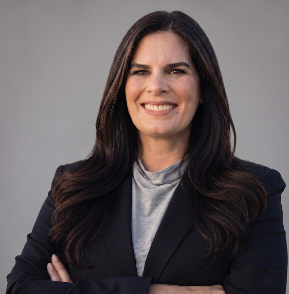A woman with long dark hair is standing with arms crossed, wearing a black blazer and light gray top, smiling at the camera against a plain gray background.