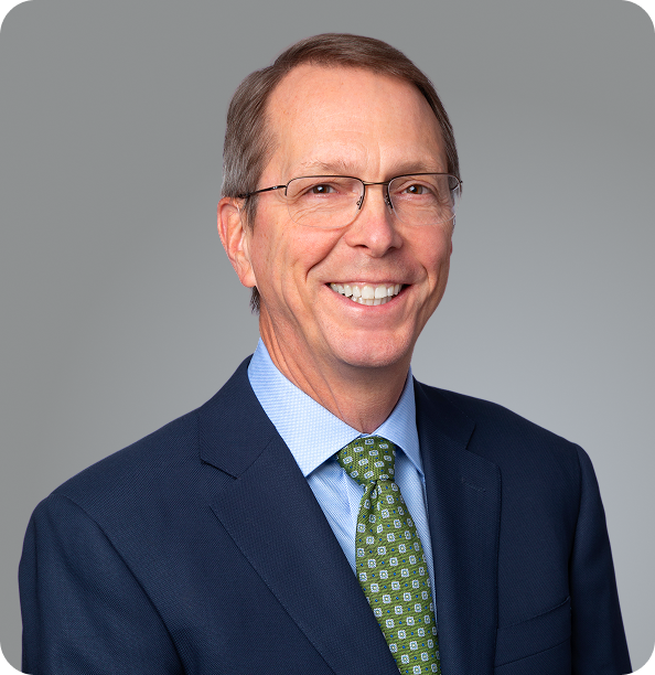 A man wearing glasses, a navy blue suit, a light blue shirt, and a green patterned tie smiles at the camera in front of a plain gray background.