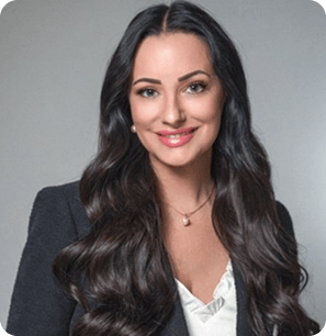 A woman with long, wavy dark hair wearing a black blazer over a white blouse, smiling at the camera against a plain gray background.