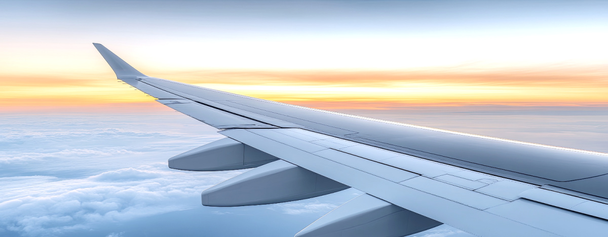 View from an airplane window showing the aircraft’s wing above clouds with a colorful sunset or sunrise sky in the background.