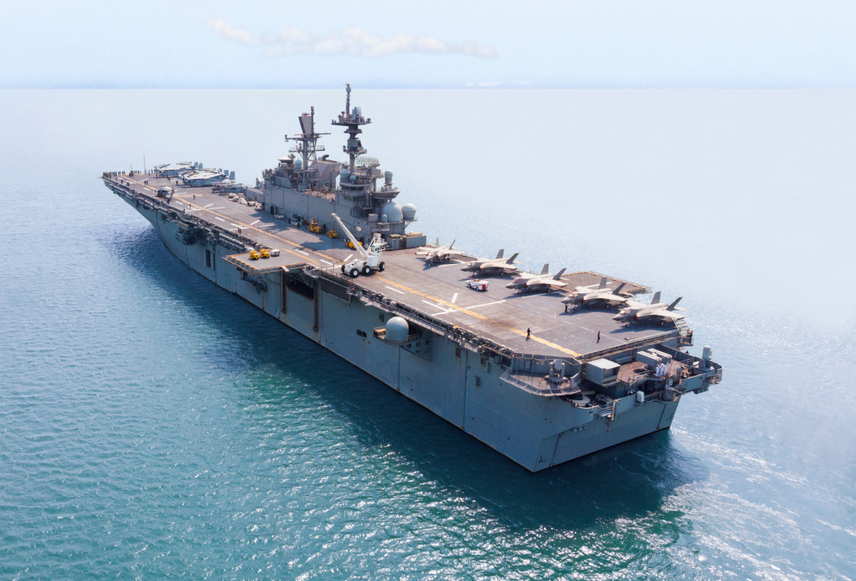 A large aircraft carrier is positioned in calm blue water, with several fighter jets and helicopters on its deck under a partly cloudy sky.