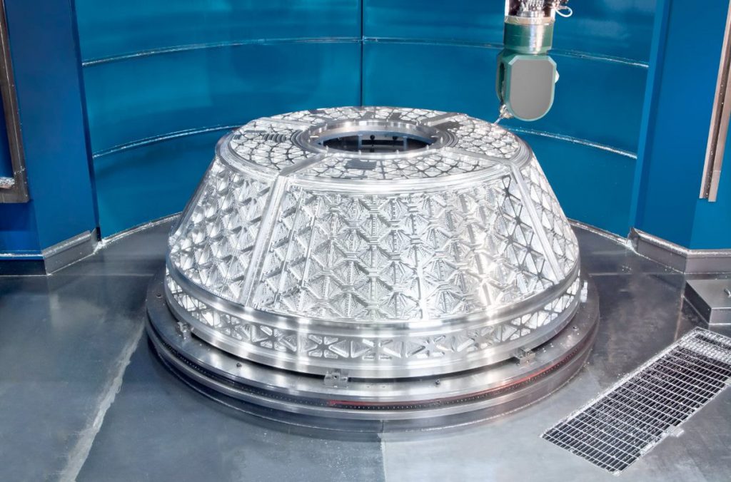 A large, precisely machined metallic component with a geometric, textured surface sits on a circular platform in a clean, industrial environment. A piece of automated machinery is positioned above the component.