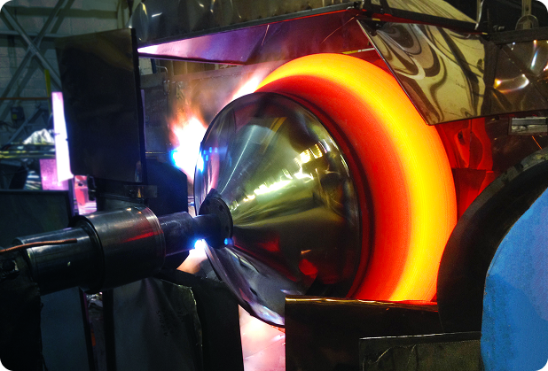 A metal spinning process in progress, showing a glowing hot metal cone being shaped on a lathe with an industrial tool in a factory setting—a vital technique for the aerospace and defense industry. The metal is bright orange due to high heat.
