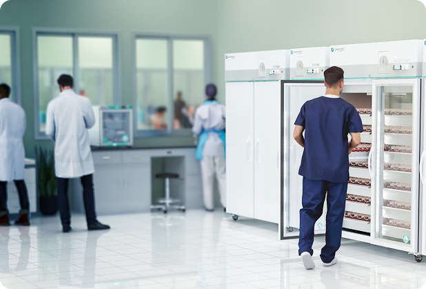 A man in scrubs stands at an open laboratory refrigerator filled with trays. Two people in lab coats work at a counter, while another conducts scientific tasks nearby. The lab has large windows and white tile floors.