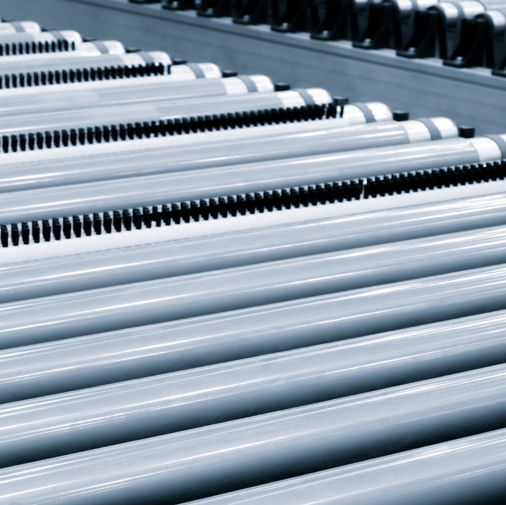 Close-up view of several parallel metal conveyor rollers arranged in rows, with a small section of black gears visible in the background, suggesting an industrial or manufacturing setting—offering a glimpse into the precision and innovation that define us. Discover more about us and our dedication to quality engineering.