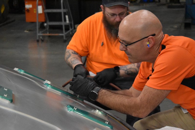 Two men wearing orange shirts and gloves work on a large metal surface in an industrial setting, attaching or inspecting green components. One man is bald and wears ear protection. The background shows industrial equipment and concrete floor.