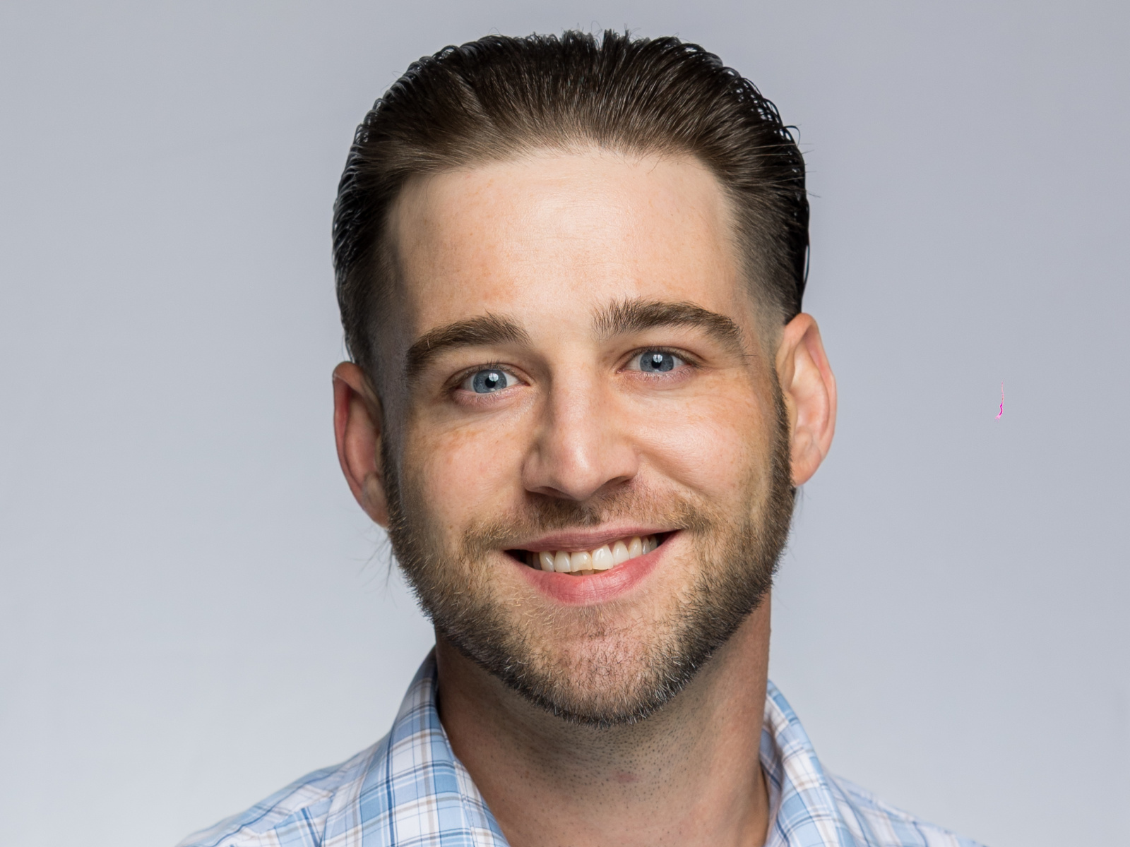A man with short brown hair, blue eyes, trimmed beard, and light skin is smiling. He is wearing a blue plaid shirt and is pictured against a plain light gray background.