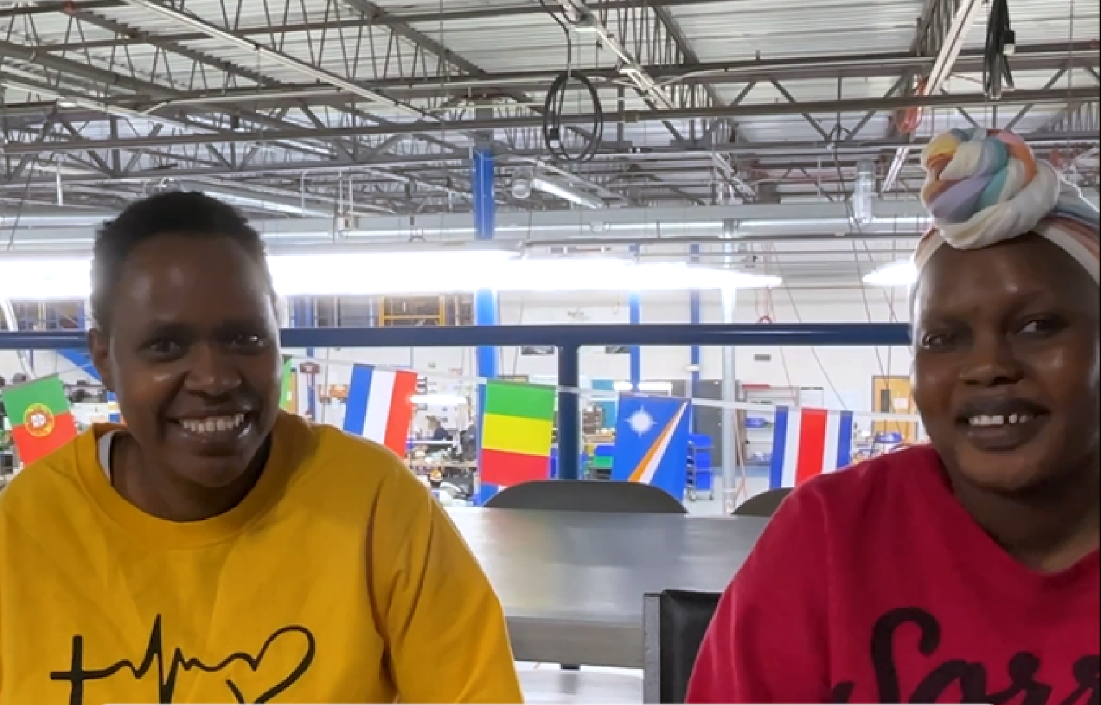Two women sit and smile in an indoor industrial space. One wears a yellow shirt, the other a pink shirt with a striped headwrap. Various international flags hang in the background above a busy workspace.