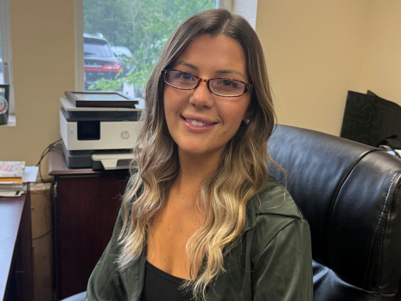 A woman with long, wavy hair and glasses sits in an office chair, smiling at the camera. There is a printer, some papers, and a window in the background showing parked cars and greenery.