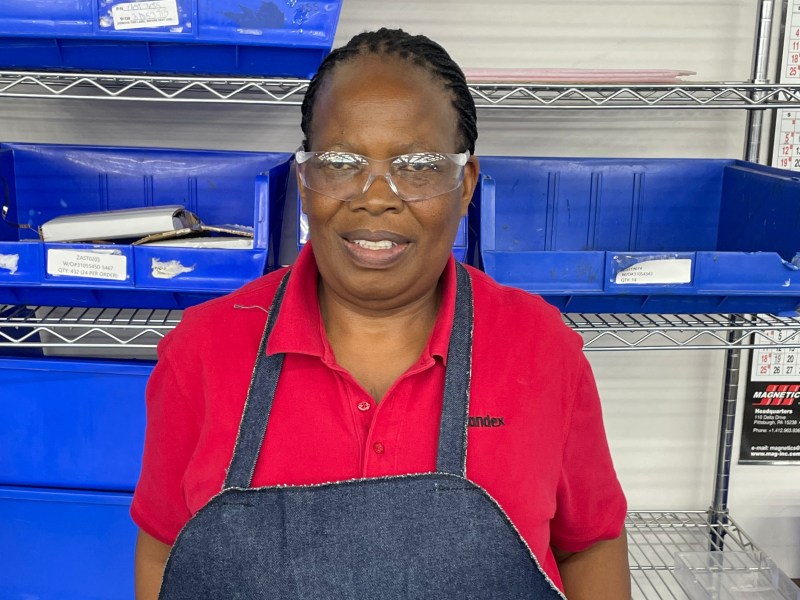 A person wearing safety glasses, a red shirt, and a dark apron stands in front of metal shelves with blue plastic bins in a workspace.