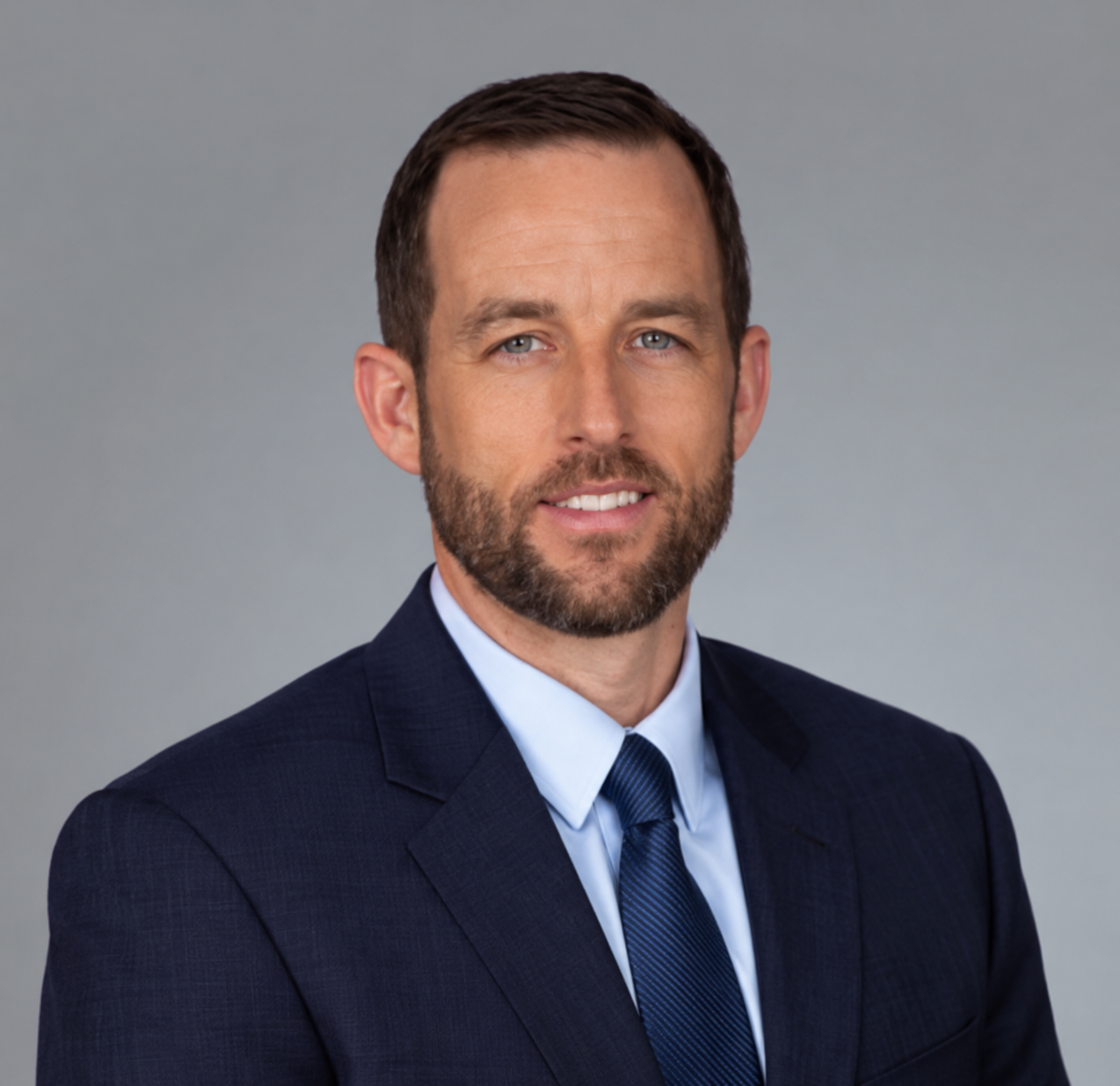 A man with short brown hair and a beard, embodying leadership, wears a dark blue suit, light blue shirt, and blue tie as he confidently poses against a plain gray background.