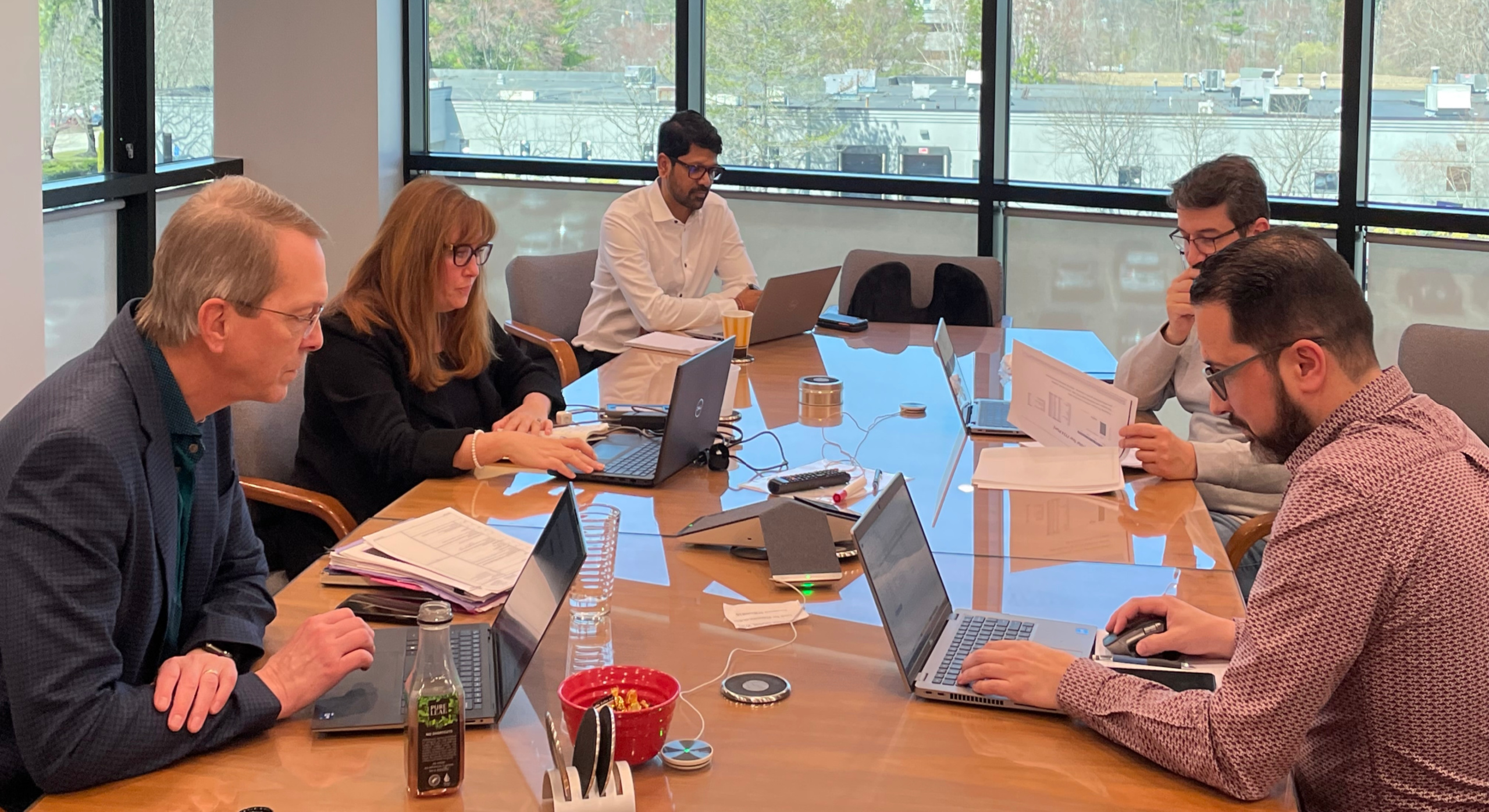 Five people sit around a conference table working on laptops and reviewing documents. The table has drinks, papers, and office supplies. Large windows show a view of trees and buildings outside.
