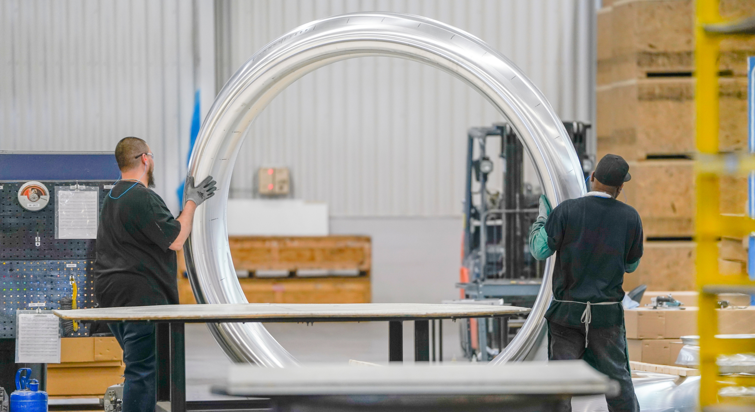 Two workers in a factory move or inspect a large, circular metal part. Industrial equipment and stacks of materials are visible in the background.