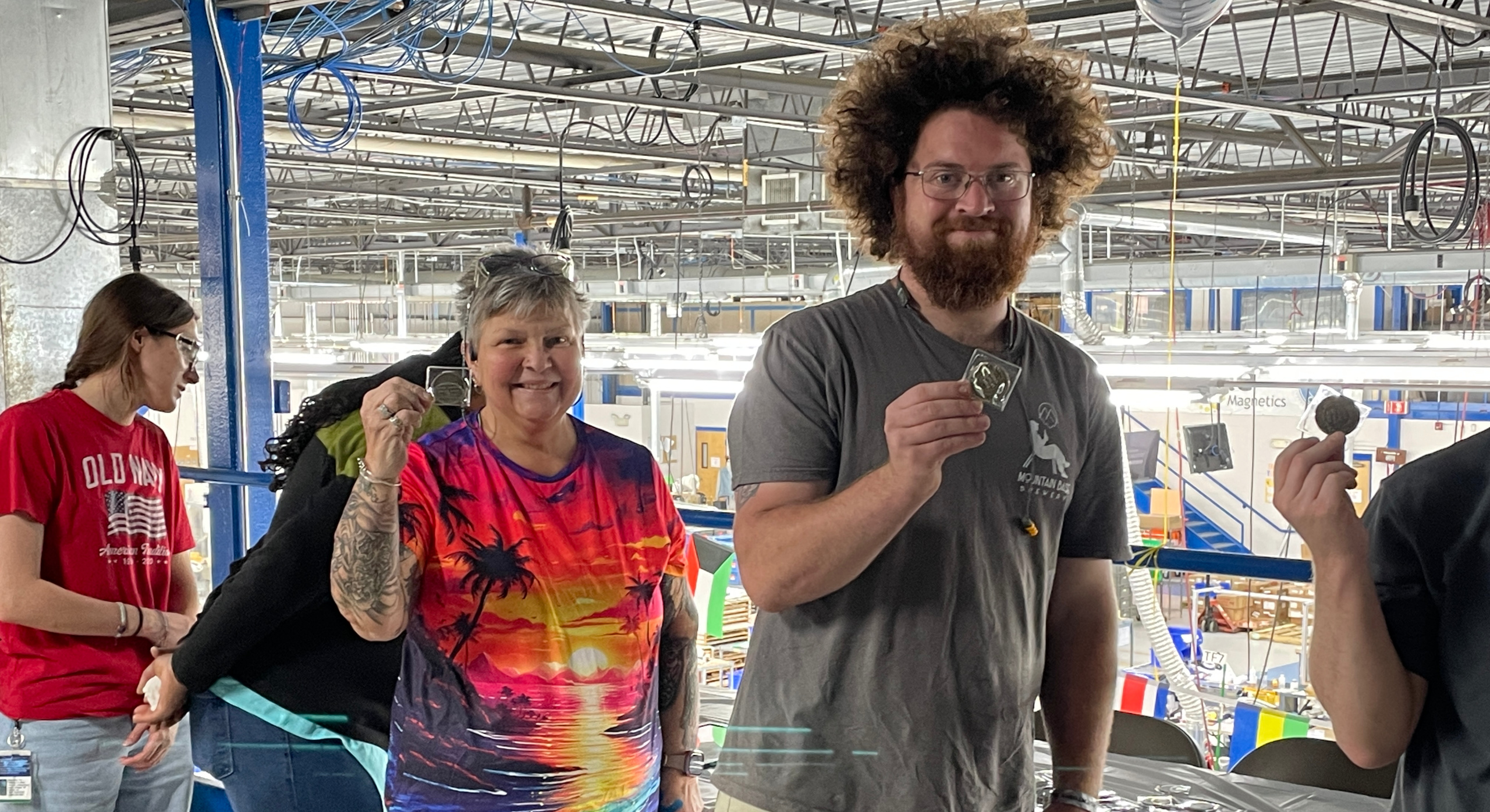 Four people stand indoors near a railing, each holding a round object. The person in front wears a colorful shirt and smiles, while the person next to them has curly hair and glasses. Factory equipment is visible in the background.