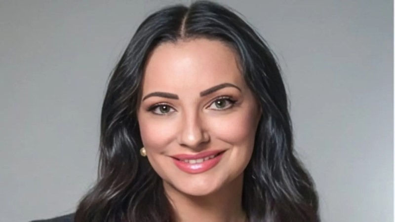 A woman with long dark hair and light skin smiles at the camera. She has brown eyes, arched eyebrows, and wears subtle makeup with pink lipstick. She is wearing pearl earrings. The background is plain gray.
