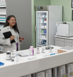 A pharmacist stands behind an Engineered Components Company counter in a pharmacy next to a tall glass-door medical refrigerator and a countertop freezer.